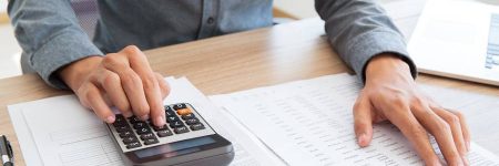 Close-up of unrecognizable accountant calculating company income and examining report. He using calculator to check financial data. Man working at table. Investment concept